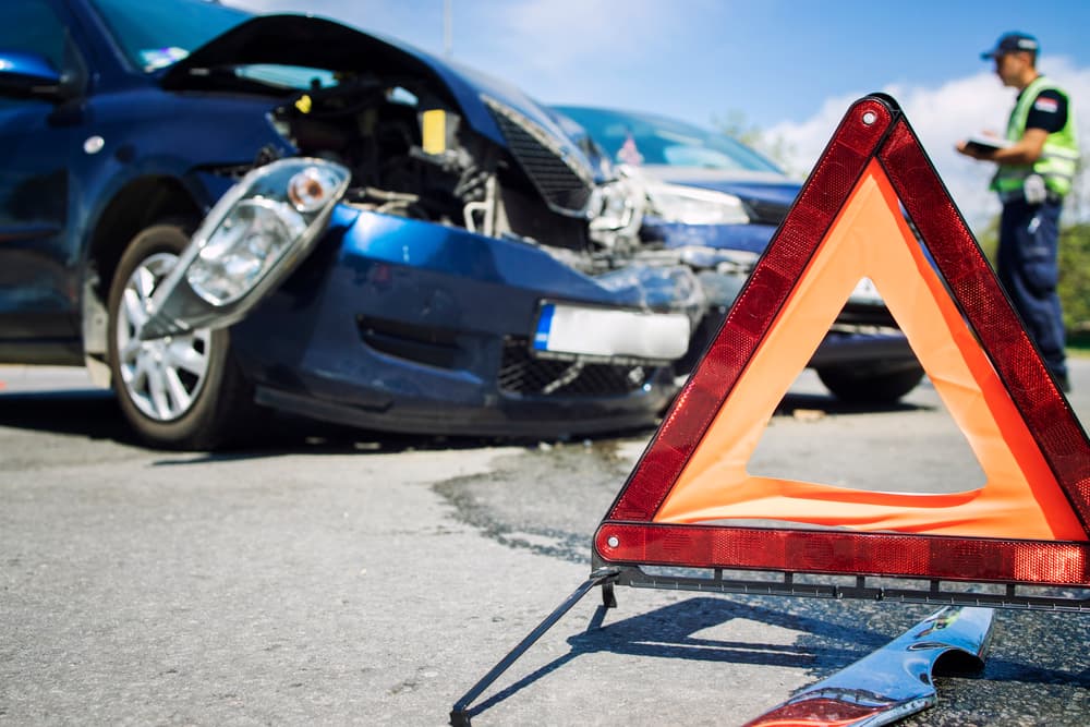 A car accident at an intersection with damaged vehicles and a warning triangle. In the background, a police officer writes an accident report.