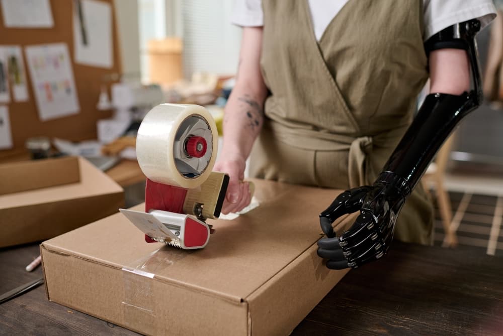 A young warehouse worker with a myoelectric prosthetic arm is sealing a cardboard box using clear packing tape.