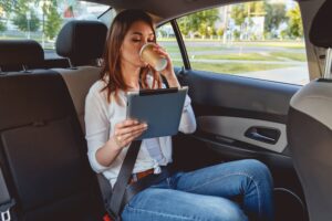 A young, attractive woman sitting in the back seat of the car, holding a tablet and drinking coffee.