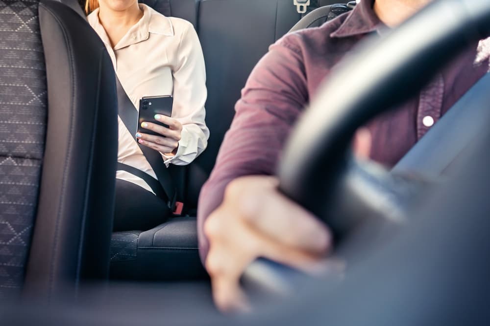 The image depicting a woman passenger using a cellphone in the back seat of a car or taxi, with the driver and another customer.