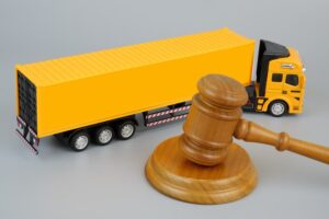 A wooden gavel and a yellow cargo truck set against a grey background, symbolizing laws and transportation.